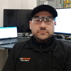 Connecting Communities participant, Luis O. He is sitting at his new job, facing into the camera with his computer behind him. He's wearing his work uniform which is a black polo that reads 