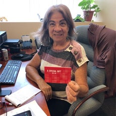 Connecting Communities Staff Member Magdalena P., holding a gift while smiling and sitting at her desk.