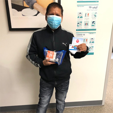 Connecting Communities participant, Norberto, posing in the Beaverton Connecting Communities office. He is wearing a protective mask, but you can tell he is smiling. He's holding some supplies provided by our program, including a gas card to help him commute for his new job. He's wearing a black Puma track jacket and jeans.