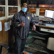 Homeless Veteran Reintegration Program participant, Cedric Graves. He is in his security officer uniform, in the security booth at his job. He's giving a thumbs up and smiling under his mask. It's night time in the photo.
