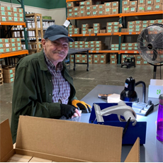 HVRP Participant, Charles is sitting at his workbench at his new job. He's smiling and wearing a blue hat, green jacket, and gloves. Location: warehouse at Precision Fulfillment