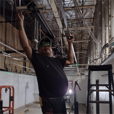 HVRP Participant, Daniel. He is at work, as a construction manager. He os wearing a baseball cap, sunglasses, and a black t-shirt. He is standing on a ladder, and working on a cross-beam above his head.