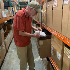 Harry in his workplace: the Precision Fulfillment warehouse. He is an older white male. He's wearing a tan hat, glasses, a red polo, and khakis. He is putting an item in a box, on a shelf.