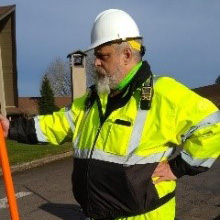Mark is wearing a neon yellow flagger uniform, holding a stop sign at his new job, as a flagger at A+ Flagging. Location: a residential street.