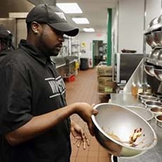 Homeless Veterans Reintegration Program participant Michael A. - He's at his new job at Wingstop, working in the kitchen. He is holding a bowl with wings and sauce in it, flipping the wings to cover them in sauce. His uniform is a black button-up and a black baseball cap that both say 