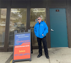 James, AKA "Patch," standing outside our Salem office. He is wearing a blue jacket, black pants, and leather shoes. He is smiling, next to a sign that reads "Proudly Serving Our Local Veterans. Easterseals Oregon Veterans Employment Program, Salem."