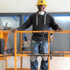 Richard, AKA RA, standing on top of a scissor lift, at his job. He is wearing a yellow hard hat, safety goggles, knee pads, jeans, a tool belt, and a grey sweatshirt.