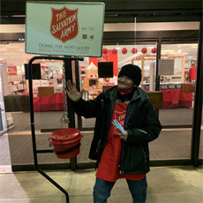 Robert is standing outside of a Macy's location. He is wearing a Salvation Army red apron, holding a bell, standing next to a donation collection stand. He is smiling and posing for the photograph.