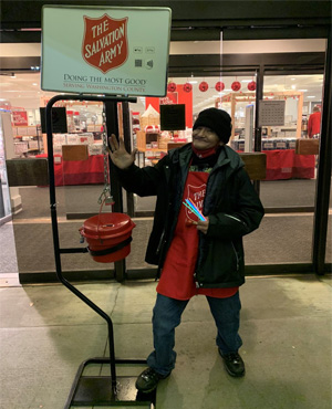 Robert is standing outside of a Macy's location. He is wearing a Salvation Army red apron, holding a bell, standing next to a donation collection stand. He is smiling and posing for the photograph.