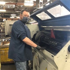 Ron is a white adult male. He has short brown hair. He is at work, in an industrial warehouse setting. He's wearing a mask, a long-sleeve blue shirt, safety gloves, and jeans.