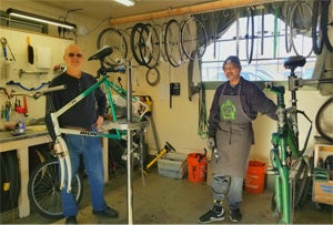 Kurt and Gabriel (employees of Sponsors, inc.) are standing inside the Recyclery bicycle repair shop, smiling for the photo. They are surrounded by bicycles, tools, and parts. Gabriel is leaning on a bicycle that he was working on.