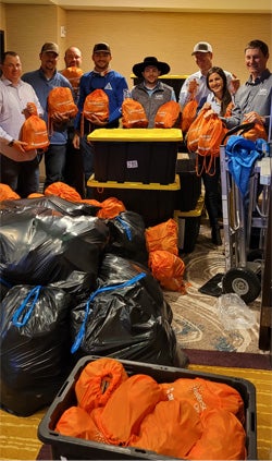 8 young men and women, holding some of the food kits they volunteered to put together. They are smiling and posing for the photograph, surrounded by numerous boxes filled with the food kits. Location: in a conference room.