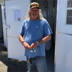 Senior Community Service Employment Program Participant, Charles Coggin. He is smiling, standing in front of a temporary housing unit, at his new job. He's wearing a hat, sunglasses, a blue t-shirt, jeans, and a watch.