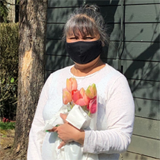 Senior Community Service Employment Program Paricipant Grace. She is standing outside, holding a bouquet of tulips, wearing a black mask, and smiling. She's wearing a white long-sleeve shirt, and her hair is tied up in a bun.