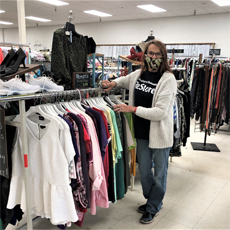 Maggie is standing near a clothing rack, inside the Habitat for Humanity thrift shop. She's sorting through clothing on hangers, and she's smiling.