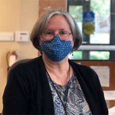 Senior Community Service Employment Program participant Rachel S. She is wearing glasses, earrings, a blue polka-dotted mask, and a black cardigan. She is sitting in our office, looking into the camera for a professional photo. She looks happy.