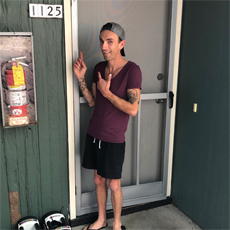 SSVF Participant B. Renfeldt, posing in front of the door to his new apartment home. He is smiling, pointing at the number for his unit. He's wearing a plum colored v-neck shirt, black shorts, a hat, and sandles.
