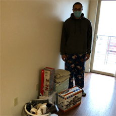 Supportive Services for Veteran Families program participant Elijah Morris. He is standing in his new apartment, wearing a mask, pajama pants, and a sweatshirt. He is standing near his collection of homegoods provided by the Homes for Heroes Foundation.