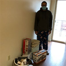 Supportive Services for Veteran Families program participant Elijah Morris. He is standing in his new apartment, wearing a mask, pajama pants, and a sweatshirt. He is standing near his collection of homegoods provided by the Homes for Heroes Foundation.