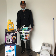 Supportive Services for Veteran Families program participant Greg Vance, posing in his new home with his Homes for Heroes welcome home kit, including a laundry basket, a vacuum, a coffee maker, and several other items. Mr. Vance is smiling.