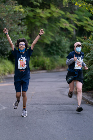 2 runners, traversing the course at the Oregon Zoo. One runner (a young man) is raising his arms in excitment and smiling. The other runner (a young woman) is giving a thumbs-up to the photographer.