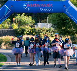 Several walkers at the live event, walking through the finish line at the Oregon Zoo. They are dressed in matching, colorful costumes that include a tutu.