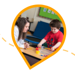 woman leans on a table watching while a boy works on a colorful activity