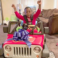 Boy smiling with arms in the air sitting in toy car with a bow on it