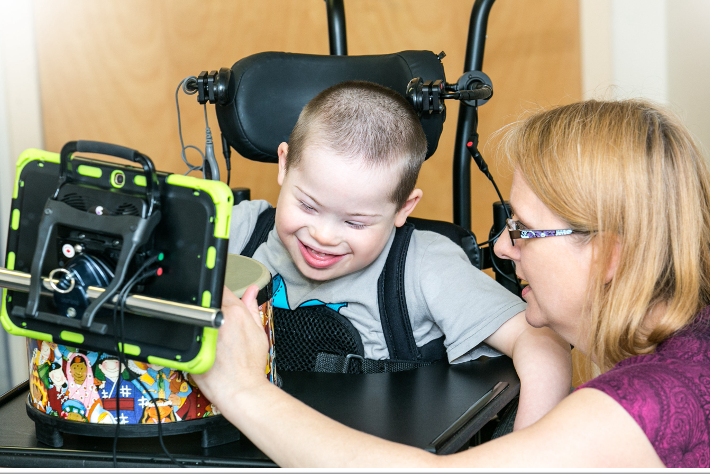 Boy in wheel chair smiling at an ipad on a tray as a woman kneels beside him pointing at something on the screen