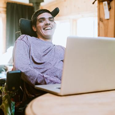 a man using a wheelchair smiles at the camera, by a desk with an open laptop