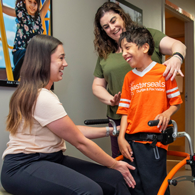 Easterseals staff smile at a young child. The child is using a walker. 