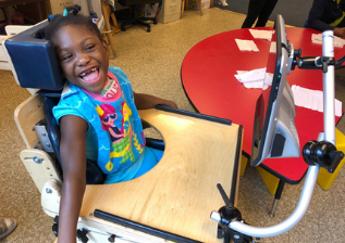 A young girl with a disability, sitting in a chair with a table and tablet in front of her, smiling at the camera