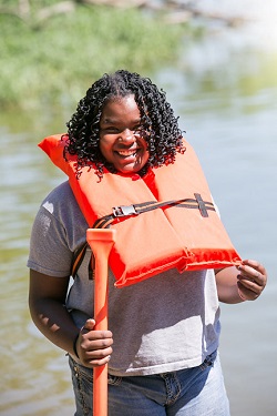 A young girl wearing a life jacket by a lake and smiling