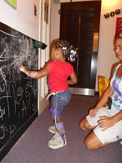 Jalea, a toddler, is standing using her assisitve devices,  playing with chalk