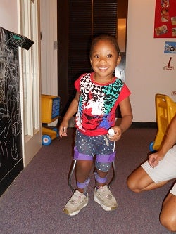 Jalea, a toddler, is standing using her assisitve devices, smiling