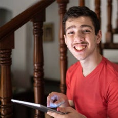 A white male in a red v-neck shirt and brown hair smiling at the camera while using his AAC tablet 