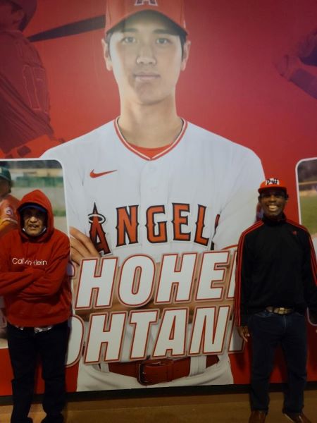 Two men standing in front of an Angel's Baseball poster