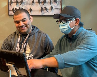 A man in a hat and mask points to an ipad to show something to the man next to him. 