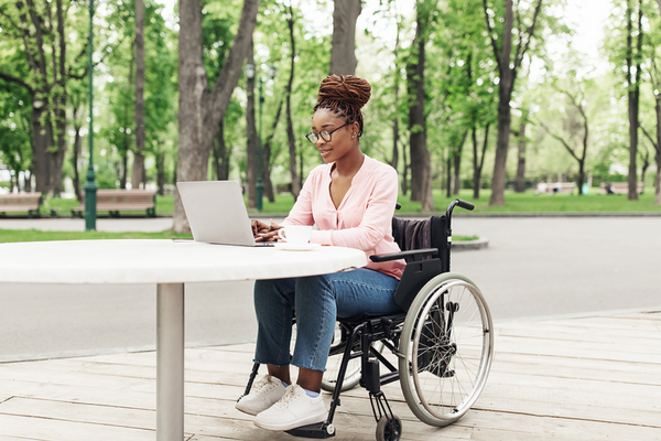 A woman in a wheelchair sits at a table outside using a laptop