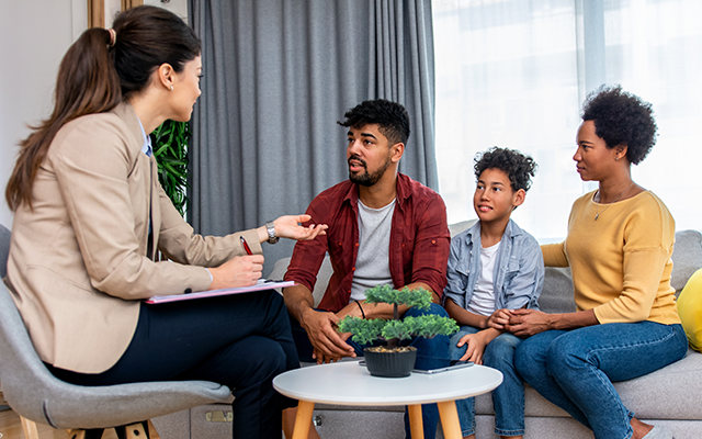 woman and man sitting on couch with kid listening to therapist speak to them while she takes notes.