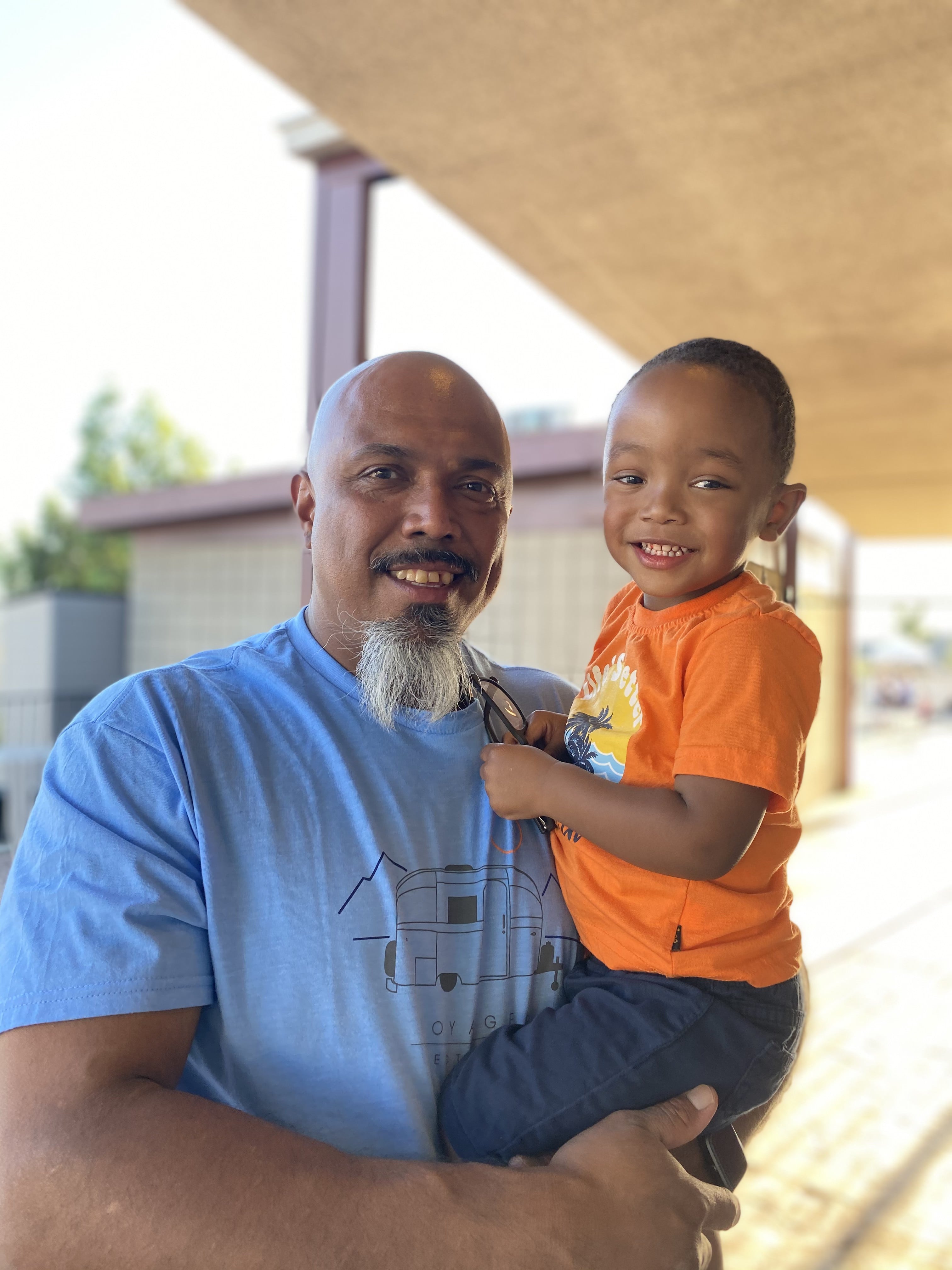 The photo shows a man in a blue shirt holding his grandson in an orange shirt. Both are smiling at the camera. 