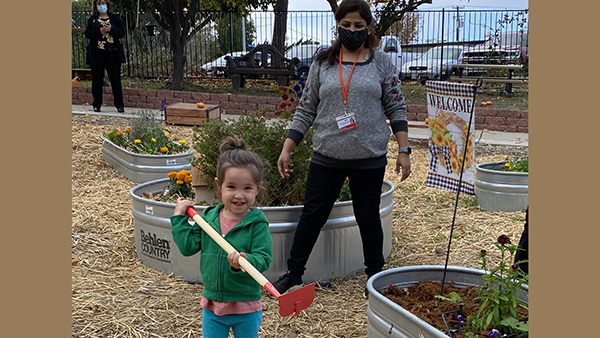 Preschool student uses a toy hoe in the garden while her teacher watches