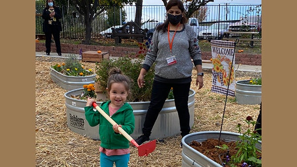 Preschool student uses a toy hoe in the garden while her teacher watches