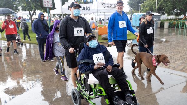 Man pushing another man in a wheelchair next to a man walking a brown dog on the Strides course in Long Beach Shoreline Village.
