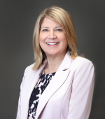 A white woman with blonder hair wearing a pink blazer and a black blouse smiling for her headshot. 