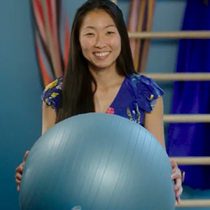 Asian woman holding up a yoga ball while smiling at camera