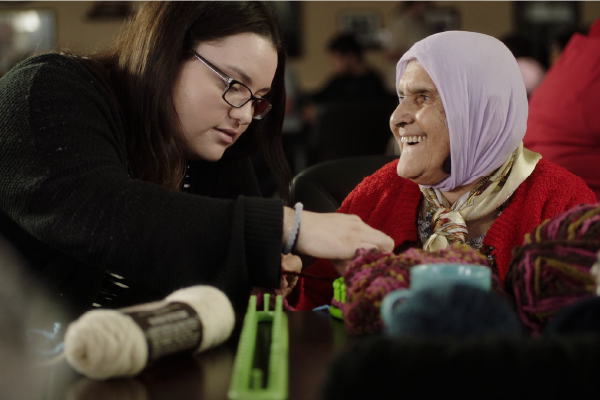 A young Hispanic woman helps older woman with a head scarf work with yarn