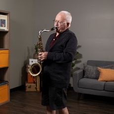 An older man wearing a sweater plays the saxophone with his eyes closed