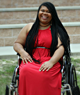Emerald sits in her wheelchair, which is on a grassy surface, wearing a vibrant red dress and smiling