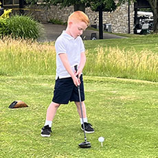Boy with red hair in a white shirt and navy shorts prepares to tee off